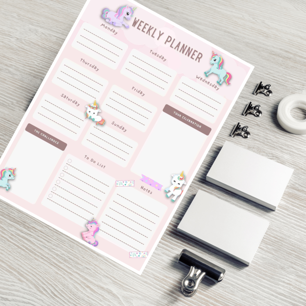 desk with clips and tape with a weekly planner that is soft pink background with purple teal and white unicorns for kids and adults.