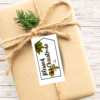 wood background, Kraft paper wrapped box with twine and a spring of evergreen, with a tag shaped tag with Blessed Christmas in black with evergreen in the corners with bells and stars