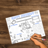 wood table with placemat of the train birthday party favor with hand with pencil ready to fill it out. Placemat shows maze, scramble, word search, how many, and tic-tac-toe. Design your train station, train tracks to fill in name to customize.