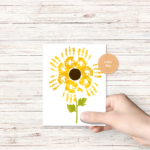 white wood background, has a hand holding the finished product of yellow handprints making the petals of a sunflower, with green stem, orange circle that says letter size