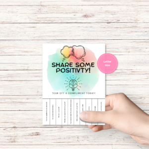 wood background, shows hand holding flyer ready to put up Positivity Compliment tear off flyer. Pink Circle Letter Size.