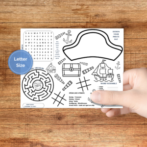 brown table with hand holding the pirate activity placemat with design your pirate hat, word search, maze, looking glass count, tic tac toe, word scramble, and speak like a pirate, and you can color the images too.Blue circle LETTER SIZE